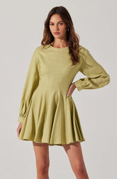 A woman is wearing a light green, long-sleeved dress with a fitted bodice and a flared, pleated skirt. The dress features slight gathering at the sleeves' cuffs and vertical seam details on the front.